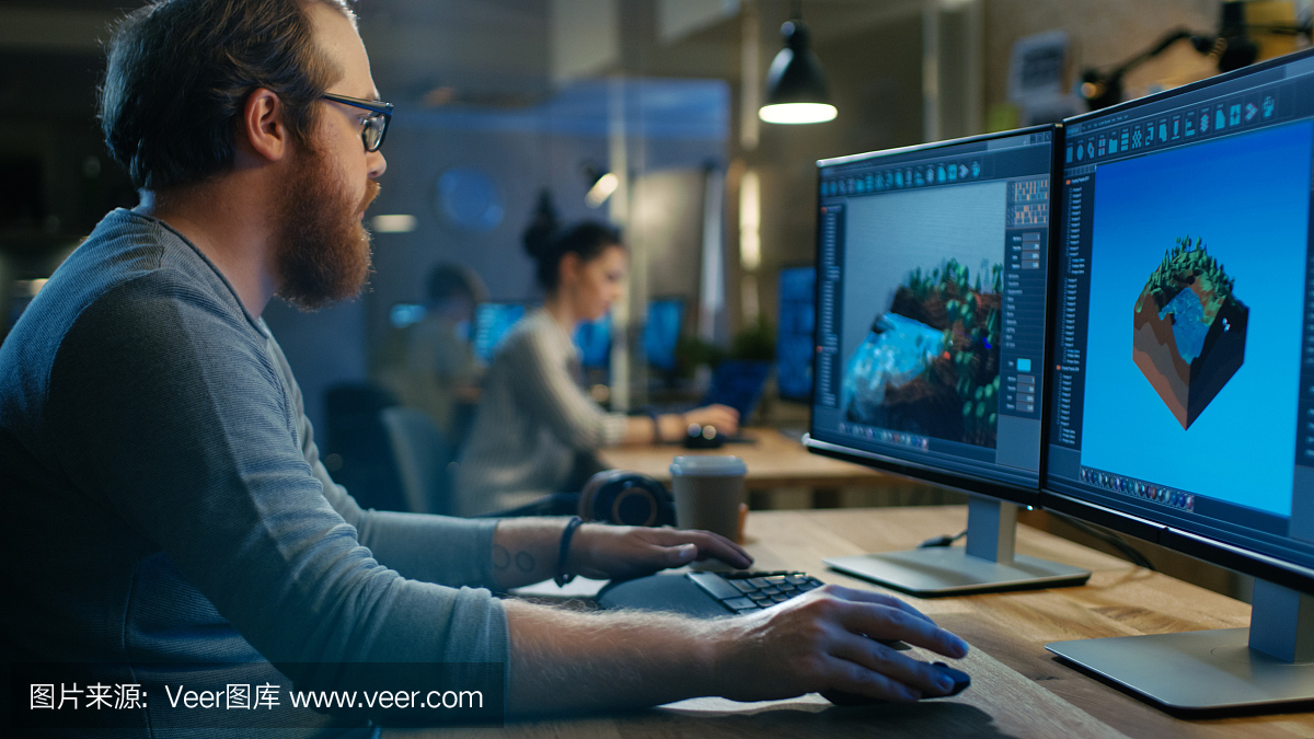 富有创造性的男性游戏开发者致力于新关卡的图像设计,他坐在自己的木桌上在两台显示个人电脑上工作。其他人在这个创意工作室工作。